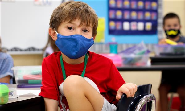boy sits at desk while wearing mask